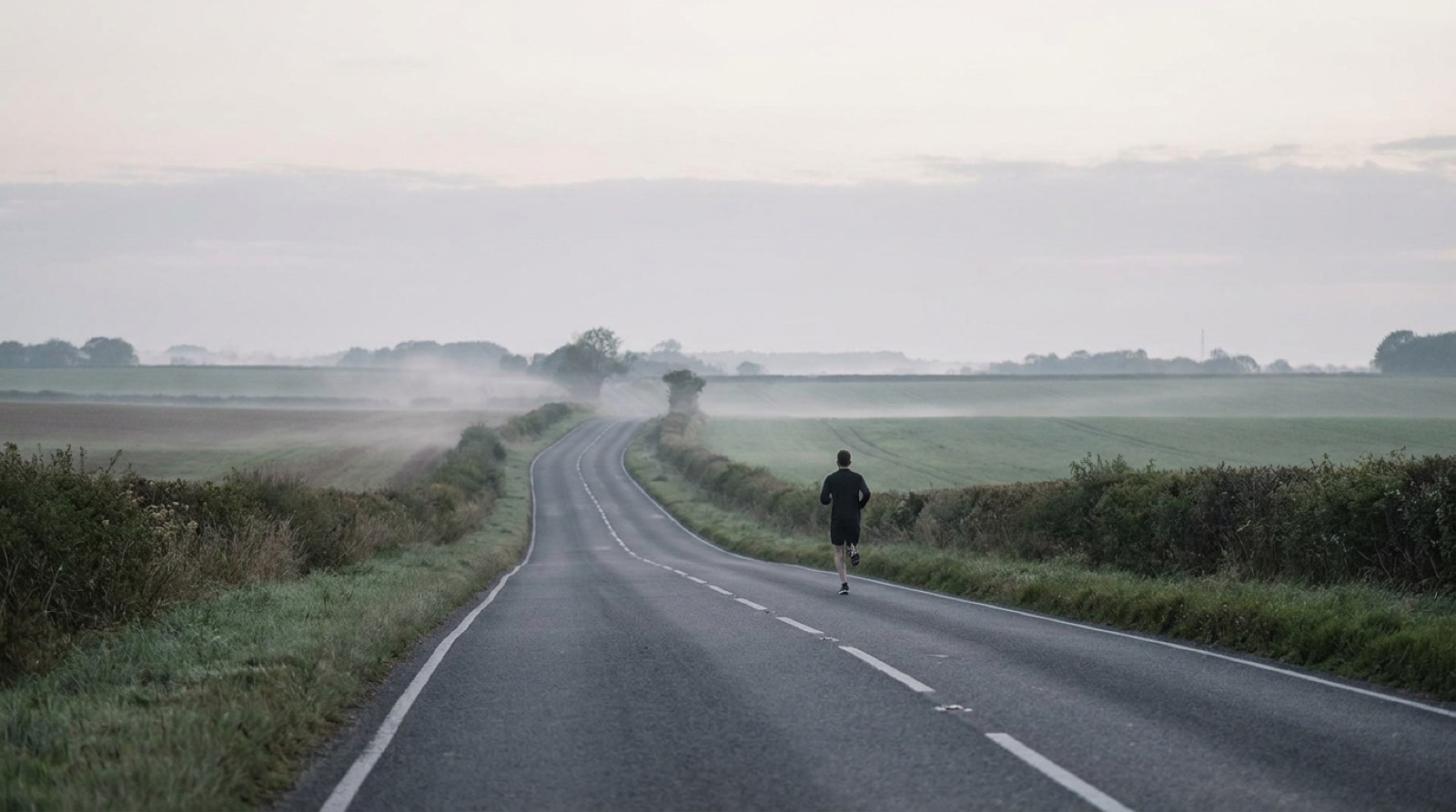 A runner on an empty open road under a soft overcast sky, seen from behind at a distance