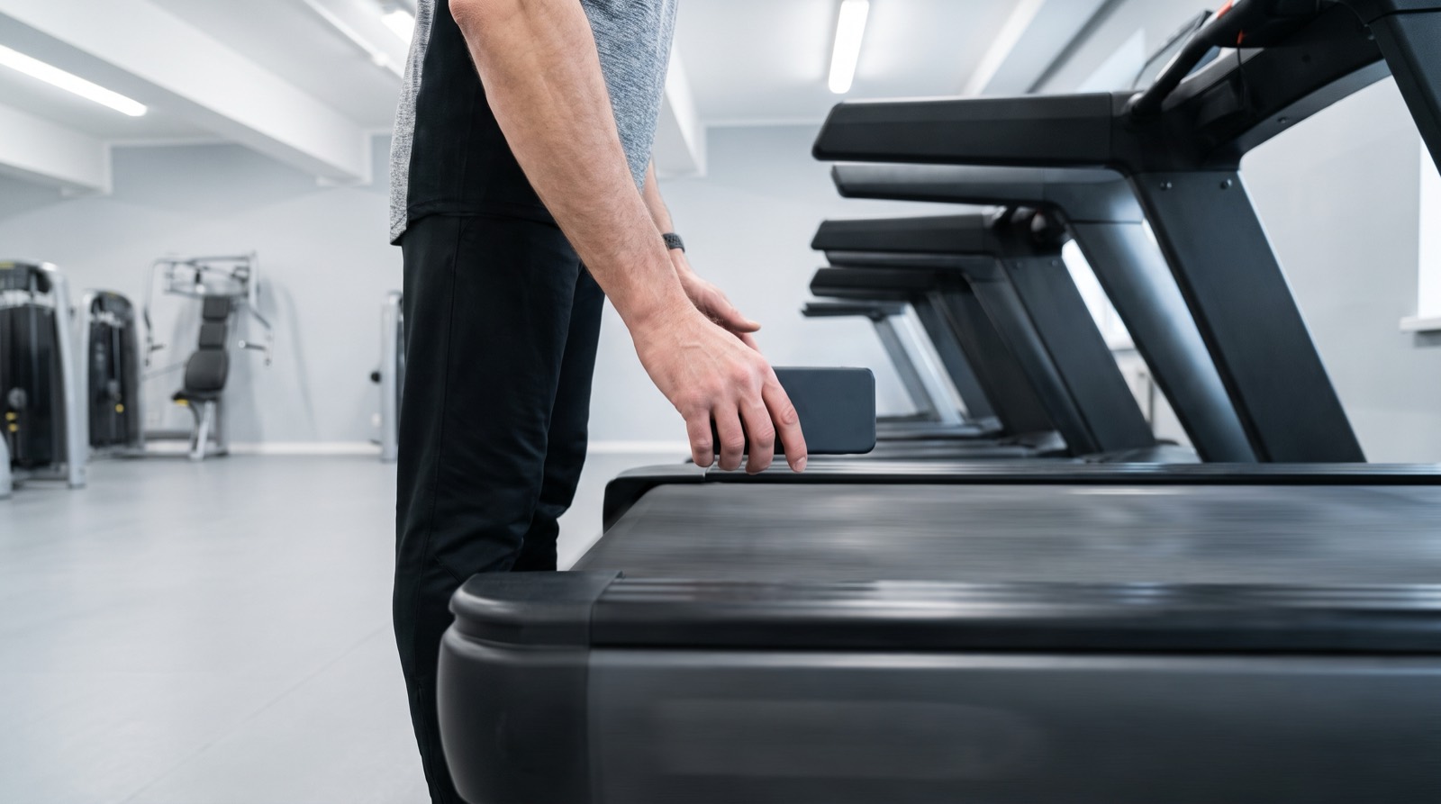 A runner standing on the floor at the side of a treadmill in a bright, plainly lit gym, holding a phone above the front edge of the moving belt