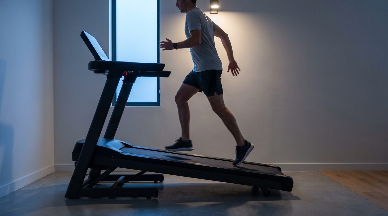 A runner climbs a treadmill raised to a steep incline, seen from the side in cool pre-dawn light