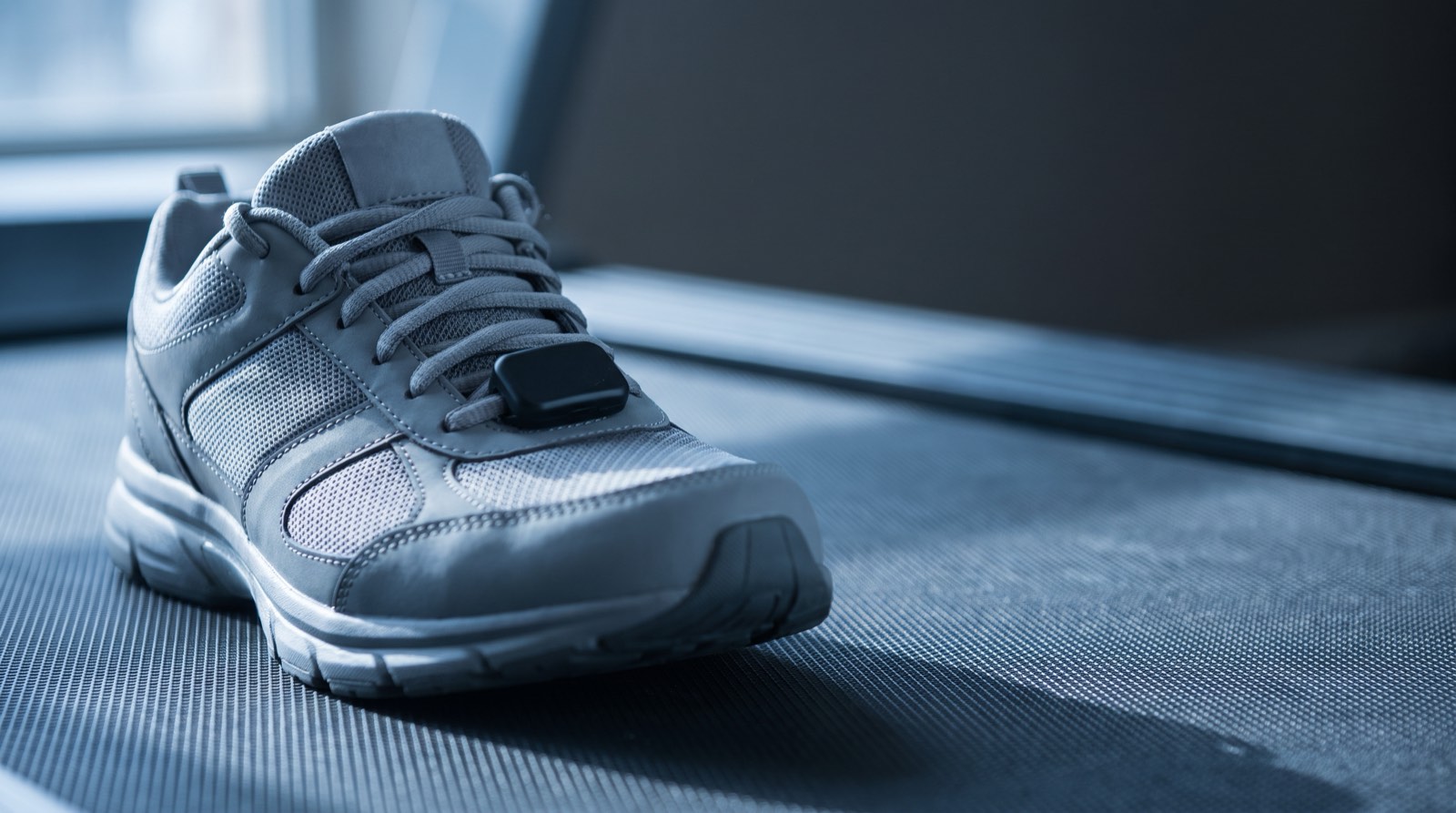 A small sensor pod clipped to the laces of a running shoe resting on a treadmill belt, lit by cool blue light before sunrise