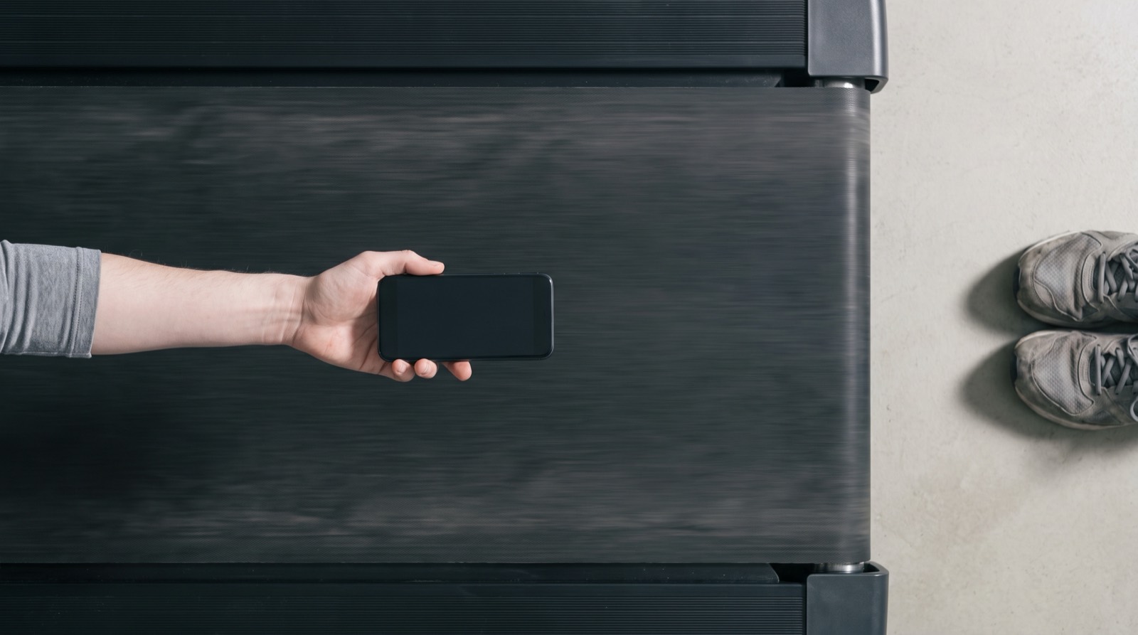 Looking down the length of a treadmill belt in soft overcast daylight, a hand entering from the left holding a phone flat above the open belt, running shoes on the floor beyond the machine