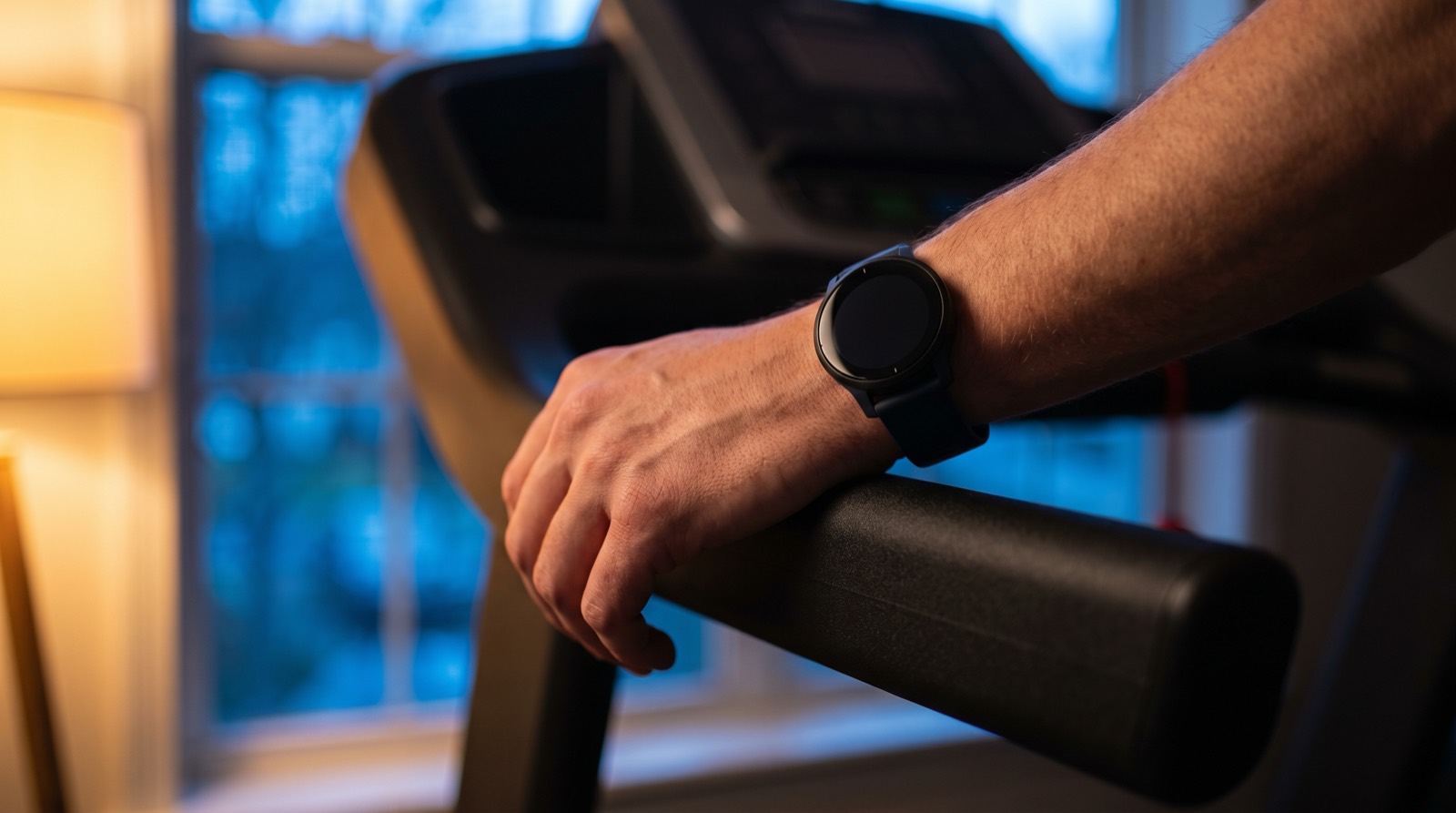 A running watch on a wrist resting along the front handrail of a treadmill in a home gym at dusk, a warm lamp on one side and cool blue evening light through a window behind