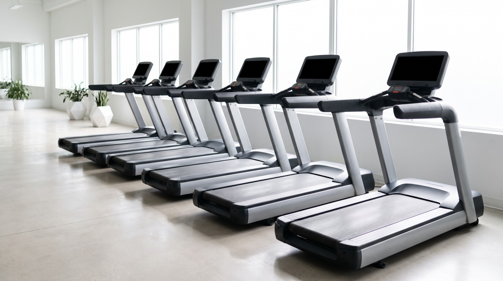 A row of treadmills standing empty and angled away in a bright white gym at midday, seen from one end of the row