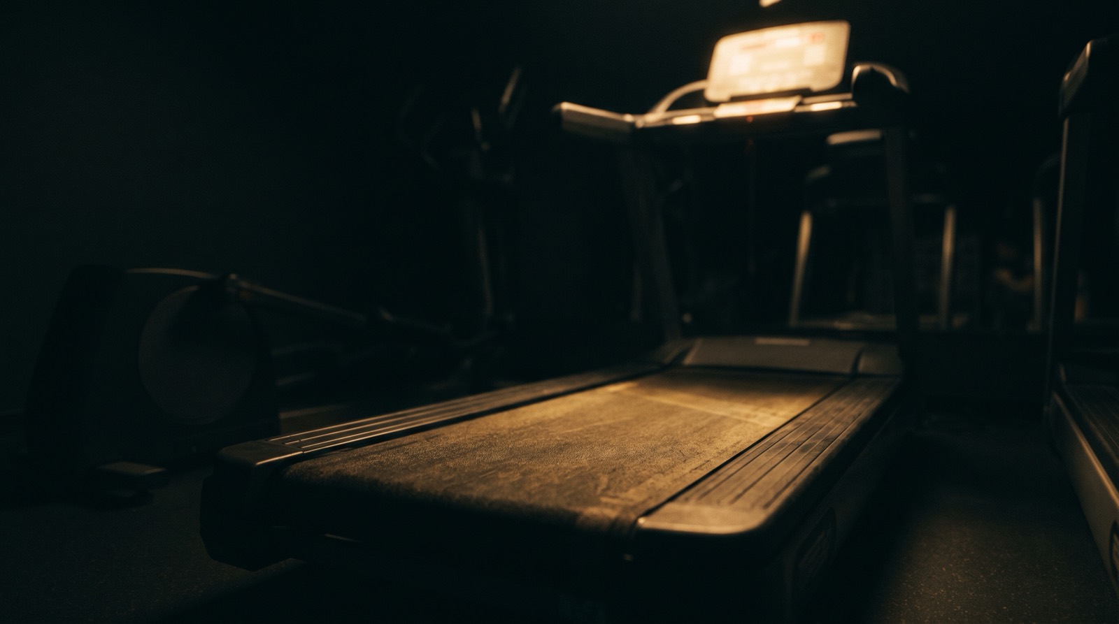 A treadmill belt in a dark gym, lit by a band of warm light, with the machine's display glowing out of focus in the background