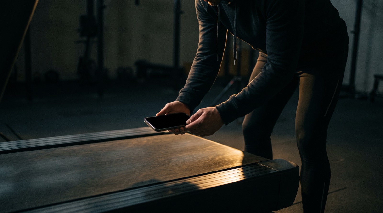Runner standing on the floor beside a treadmill, holding a phone flat and steady above the belt to measure it