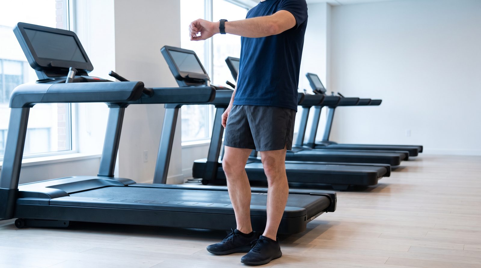 A runner steps off a stopped treadmill in a bright commercial gym at midday and lifts their wrist, the watch face hidden from view