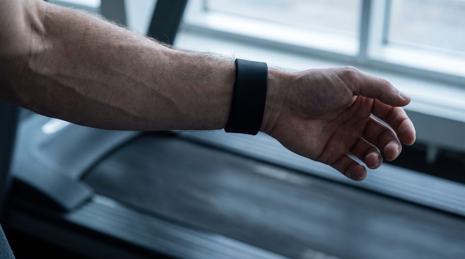 A runner's forearm viewed from underneath during a running stride, the watch face hidden against the top of the wrist, a treadmill belt blurred in the background