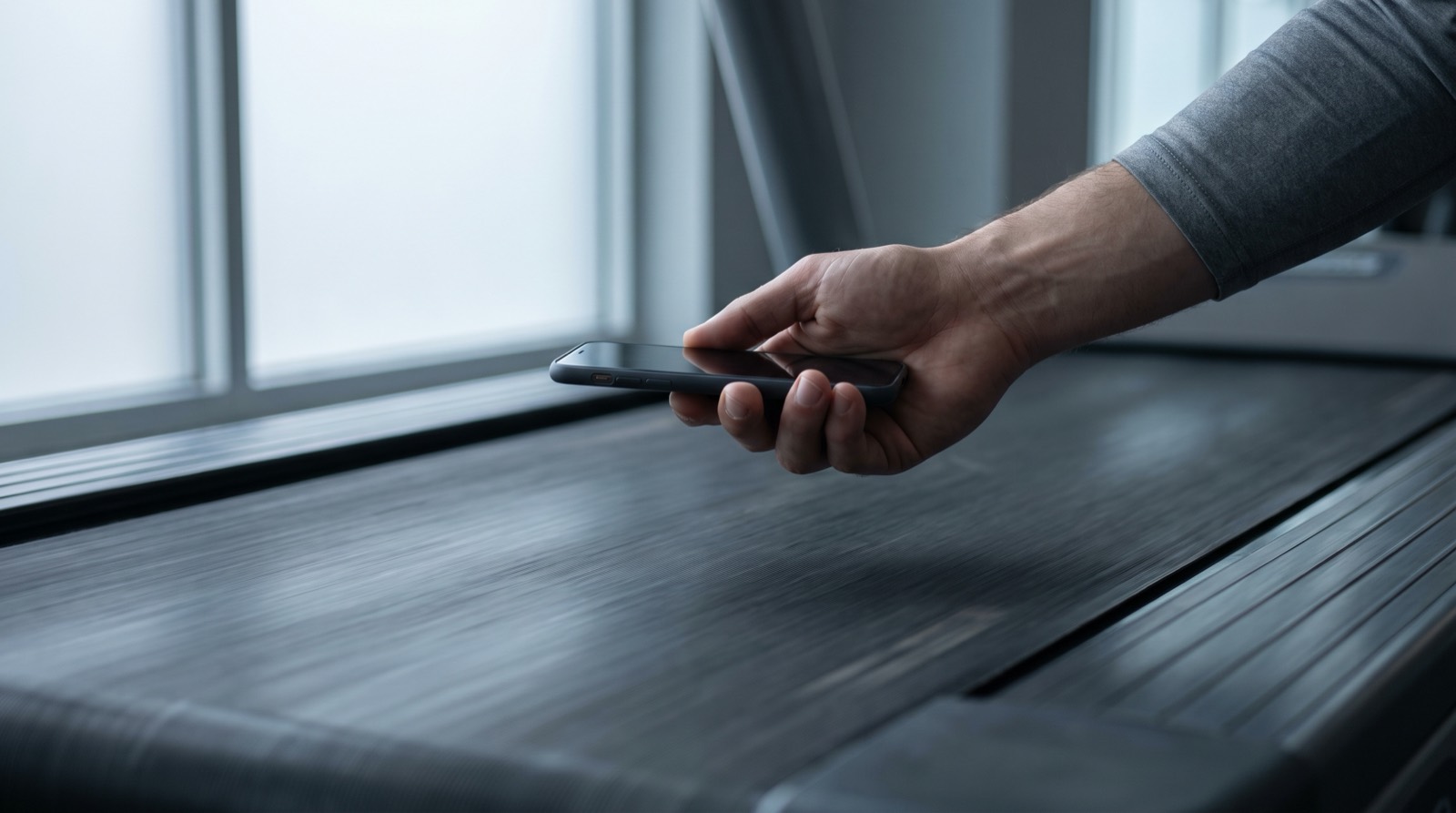 Close view of a hand holding a phone steady above a moving treadmill belt, seen from the side of the machine in soft overcast daylight