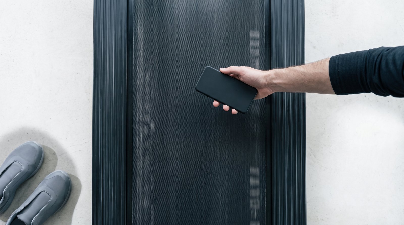 Looking straight down at a moving treadmill belt in soft overcast daylight, a hand holding a phone flat above the middle of the belt, with a pair of plain shoes resting on the floor beside it
