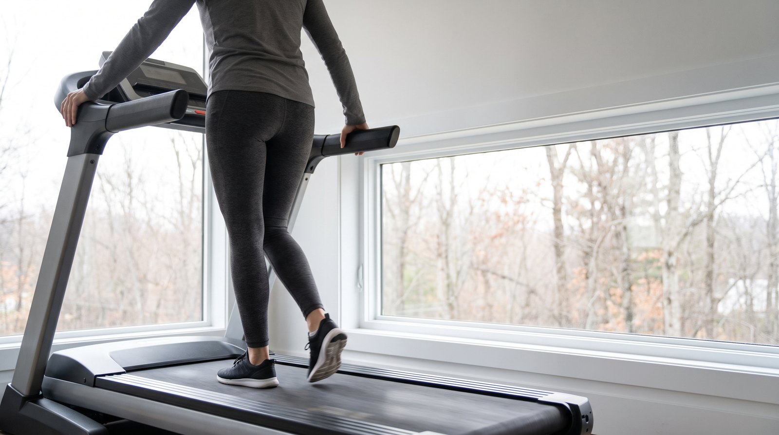 A woman at a slow walk on a home treadmill by a bright window, both hands on the side rails, her weight planted on one forward foot