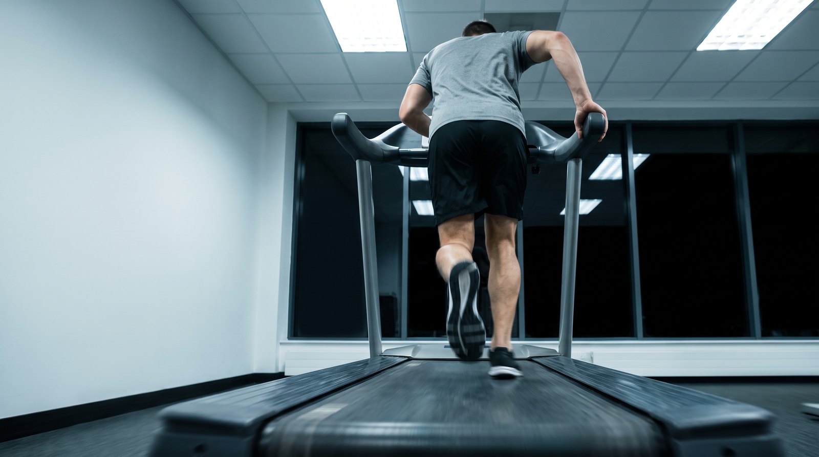 A runner seen from behind on a treadmill in a cool-lit gym at night, one hand gripping the side rail as his foot lands on the moving belt
