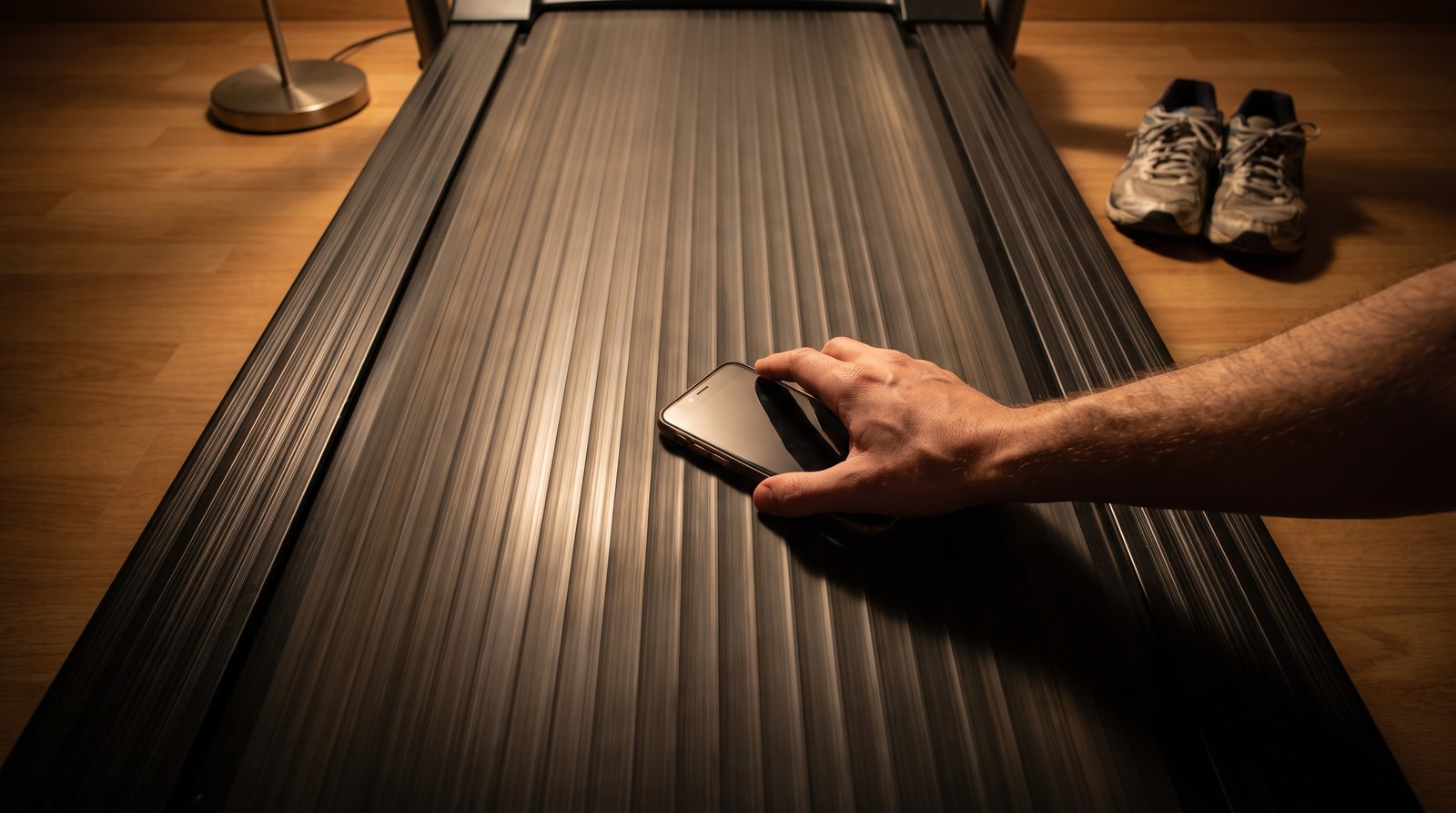 Looking down the length of a treadmill belt in warm evening lamplight, a hand entering from the right holding a phone flat above the open belt, with running shoes on the floor beyond the machine
