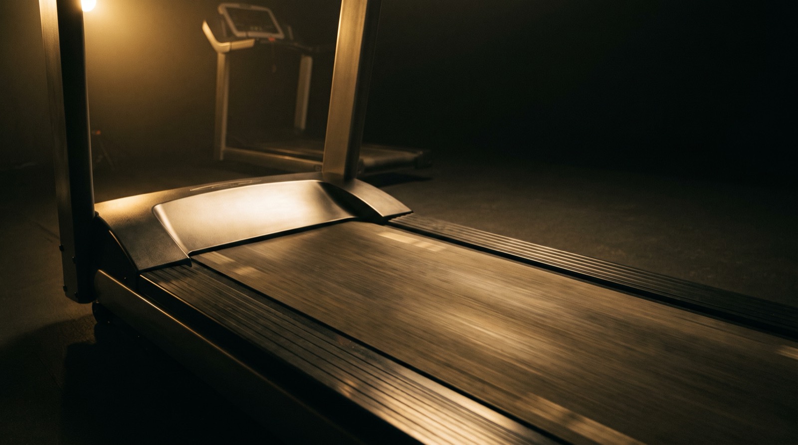 A single treadmill running alone in a dark gym at night, one warm light raking across the moving belt while the console sits out of focus behind it