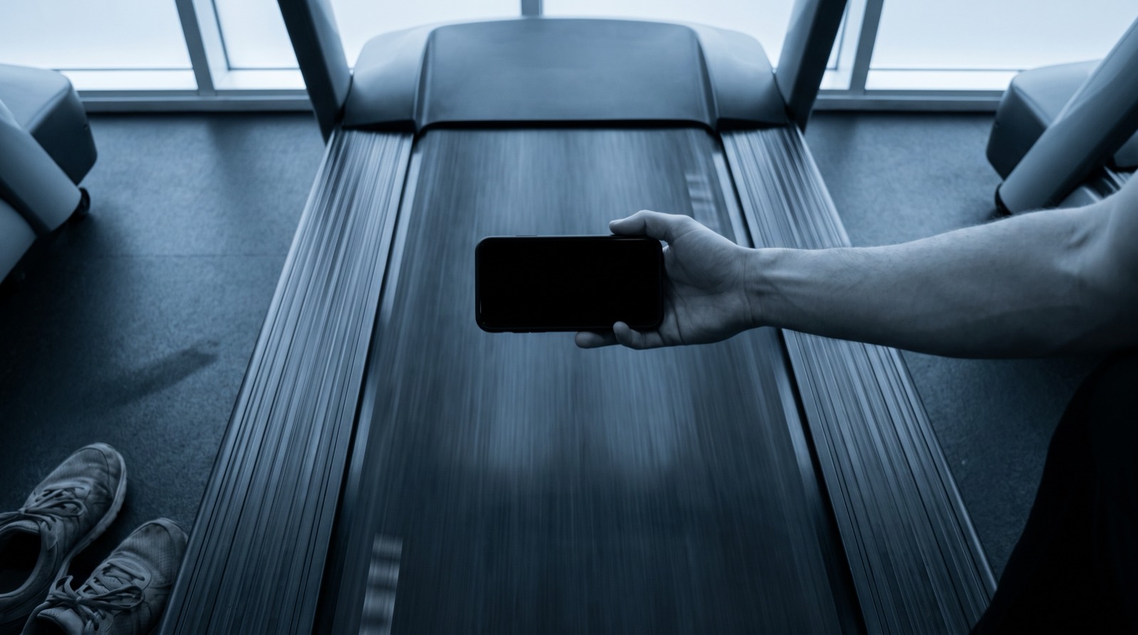 Looking down at a treadmill belt streaked with motion blur, with a hand reaching in from the right holding a phone face down above the middle of the belt
