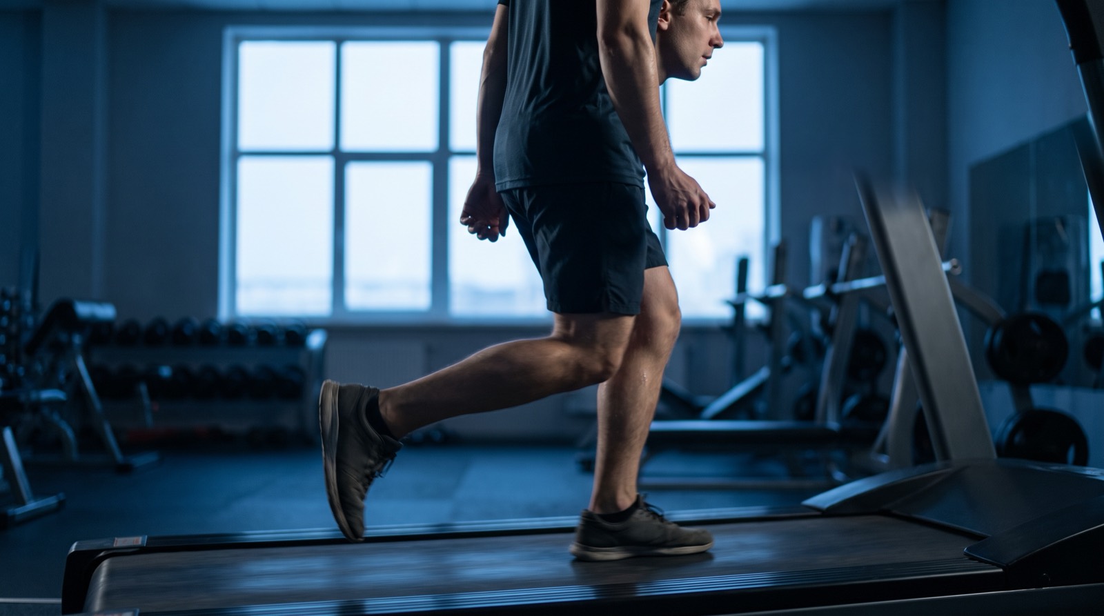 A lone runner mid-stride on a treadmill in a quiet gym, lit by cool pre-dawn blue light from a window behind, the belt blurred with motion