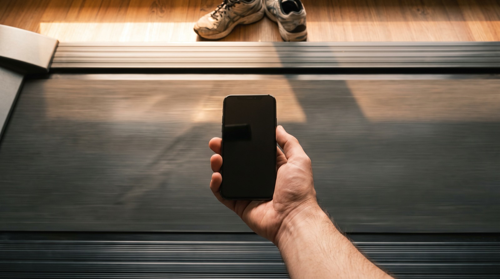 A high-angle view down a treadmill belt in warm golden-hour light, a hand holding a phone over the surface, running shoes on the floor beyond