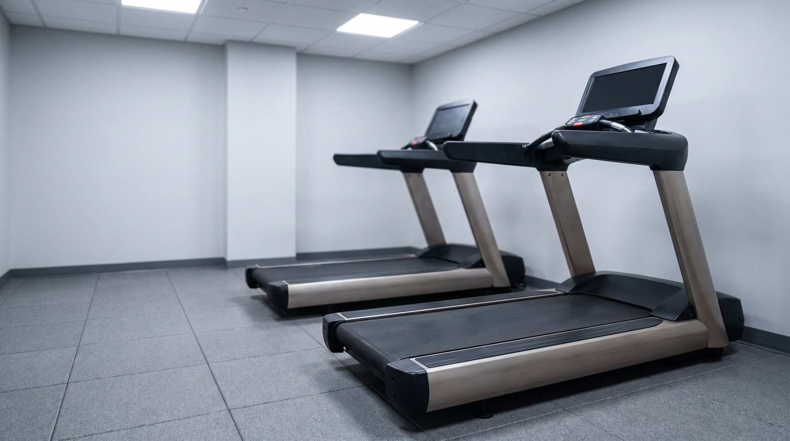 Two treadmills side by side in a plain gym room, lit evenly by overhead ceiling panels with no windows, grey rubber flooring, screens dark