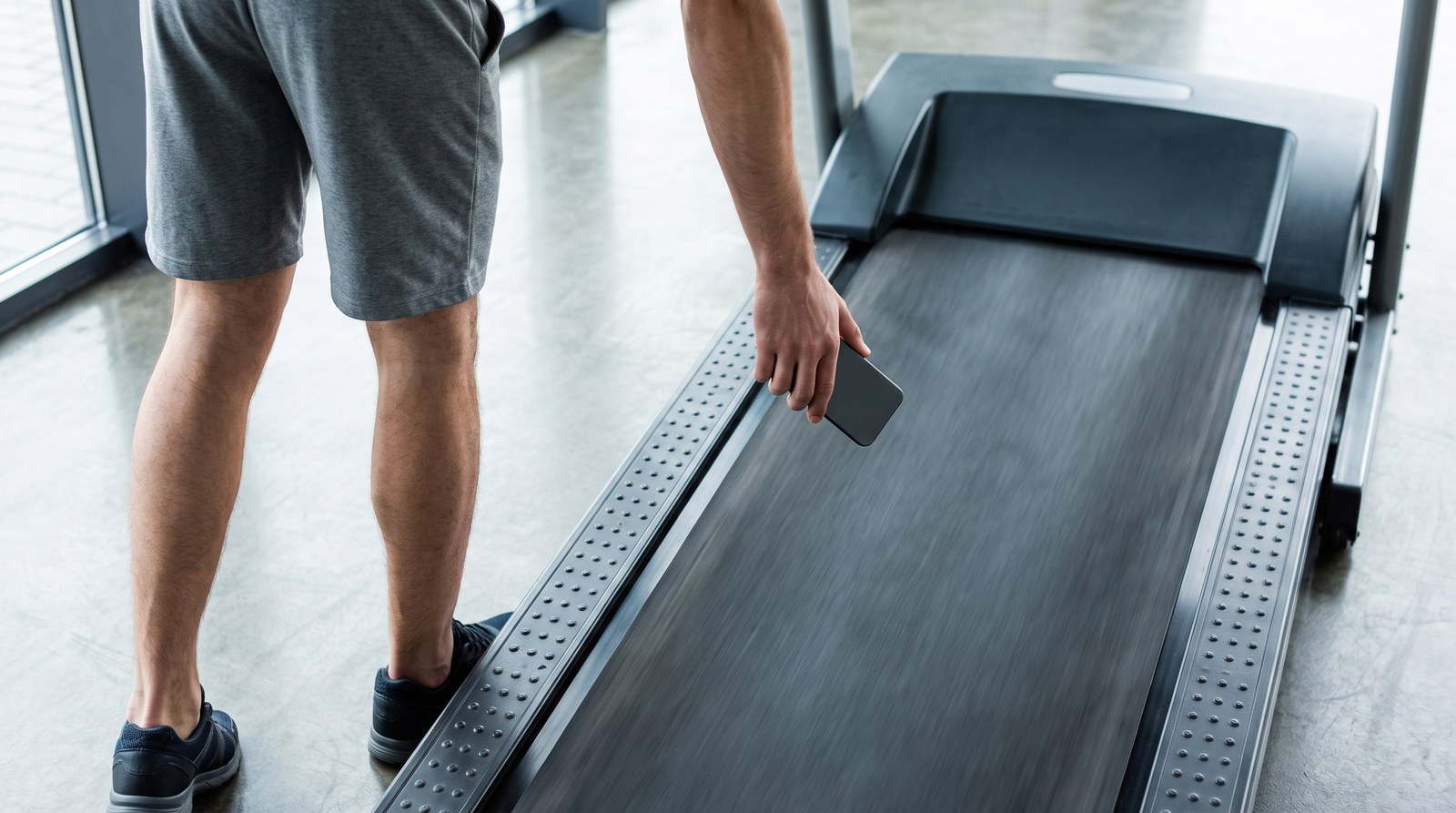 A runner standing on the floor beside a treadmill, holding a phone flat above the belt, which is blurred with motion as it runs, in bright daylight from a tall window