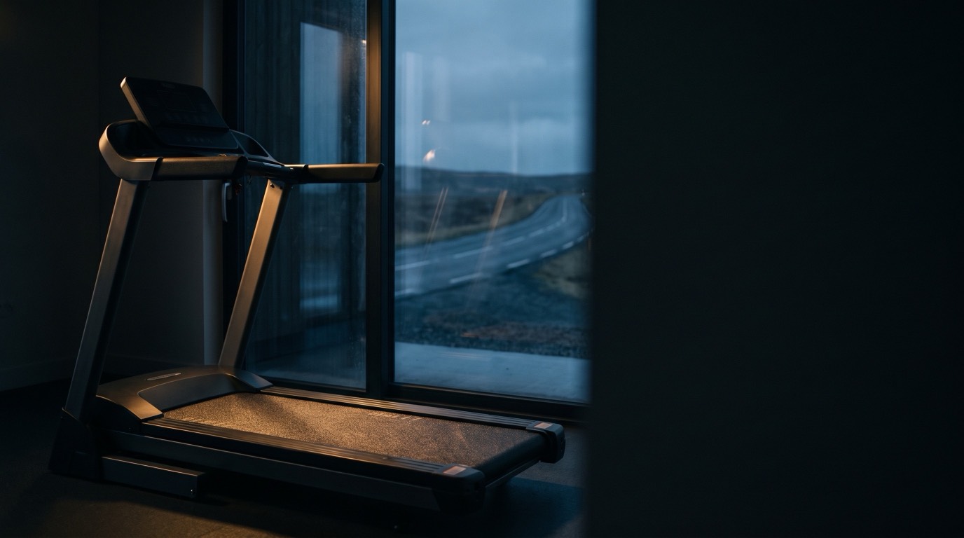 An empty treadmill standing in a dark room beside a tall window, with an open road visible outside in the pre-dawn light