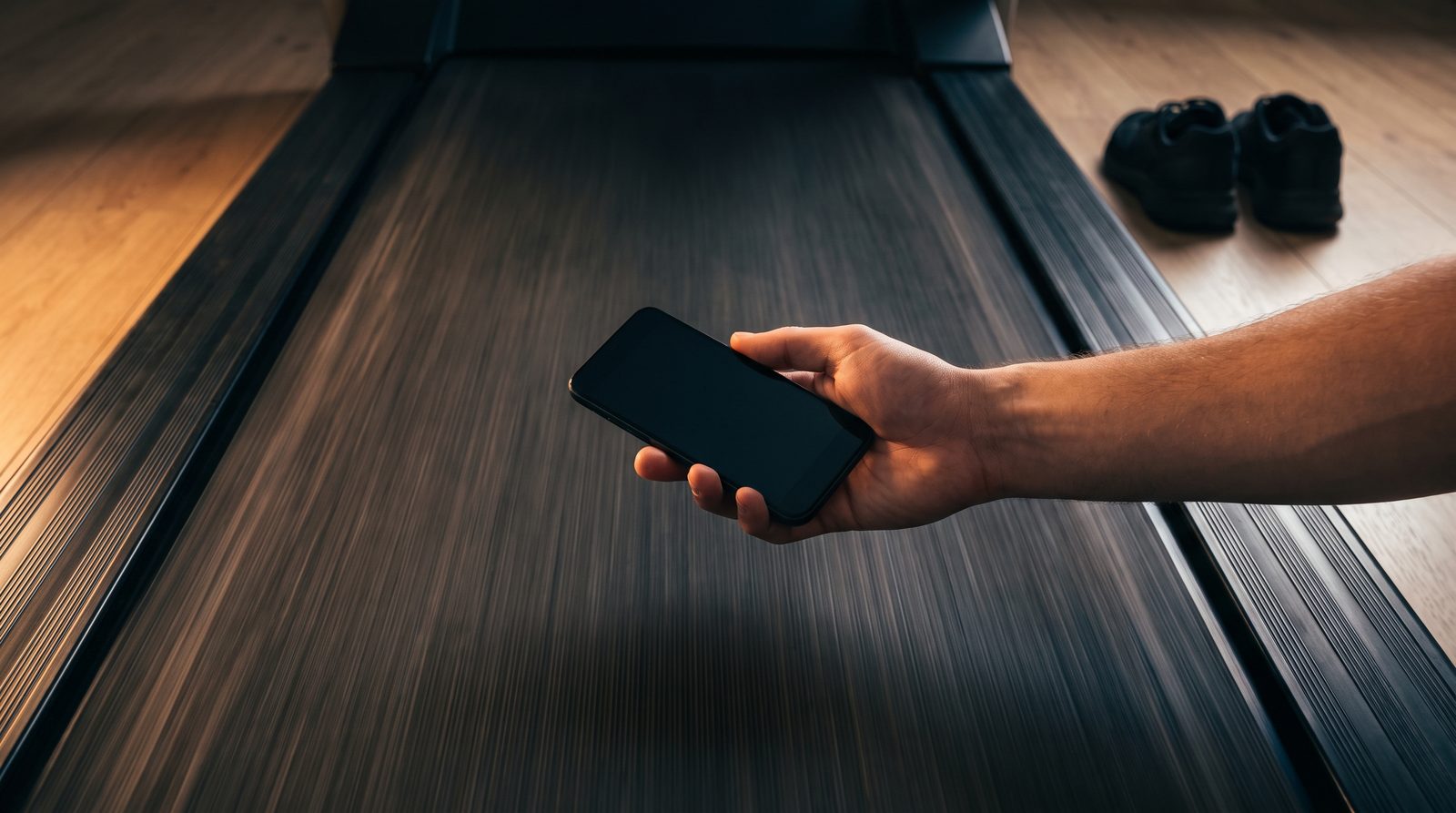 Looking down the length of a running treadmill belt in warm evening light, its tread blurred with motion, a hand entering from the right holding a phone flat above the open belt, with running shoes on the floor beyond the machine