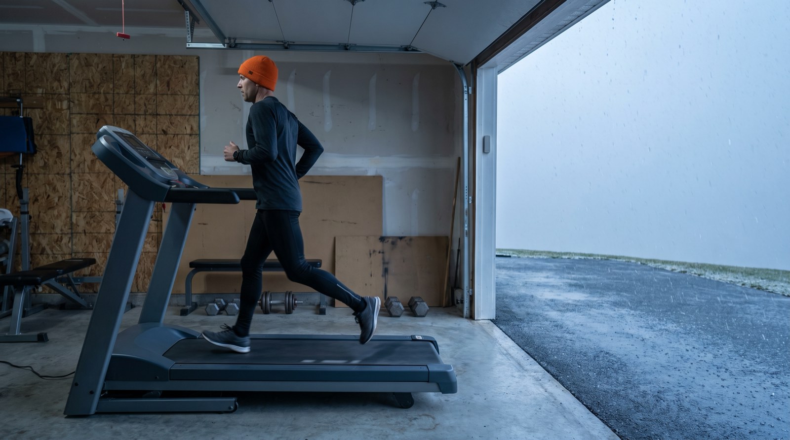 A runner in an orange knit hat runs on a treadmill in a garage with the door open to sleet on a gray late-November morning, seen from the side