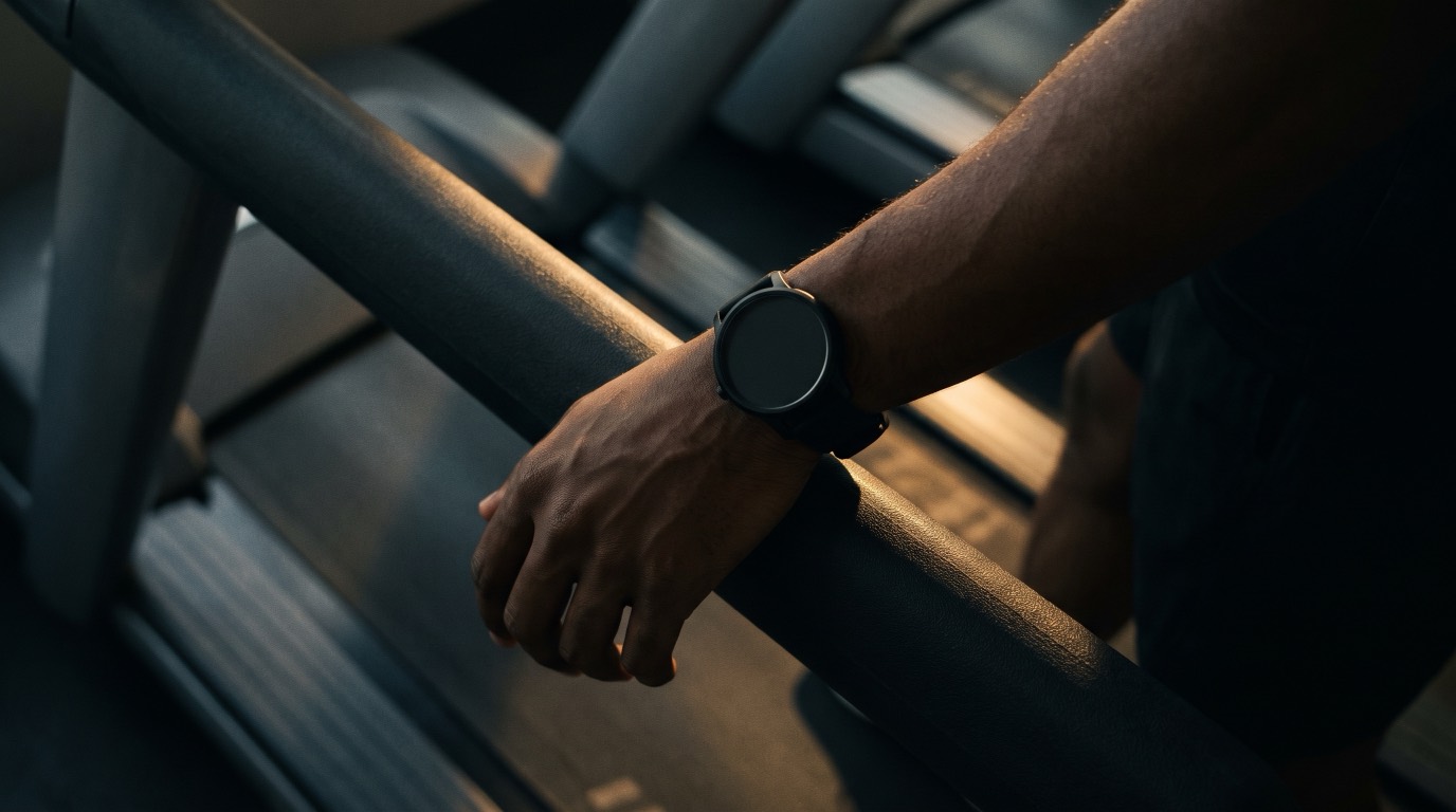 A runner's wrist wearing a sport watch, resting on the side rail of a treadmill in a dim gym lit by warm gold light