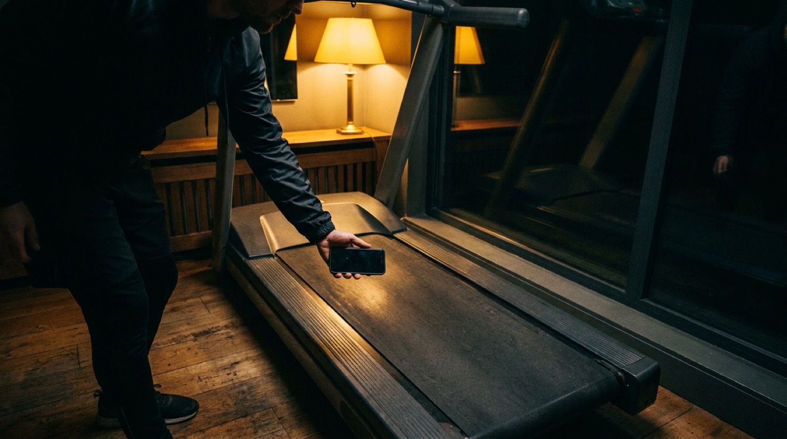 Traveler standing on the floor beside a hotel treadmill at night, reaching out to hold a phone above the belt to measure it