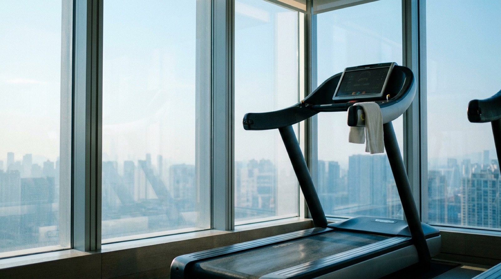 An empty hotel fitness room in the early morning, one treadmill by floor-to-ceiling windows over a hazy city skyline